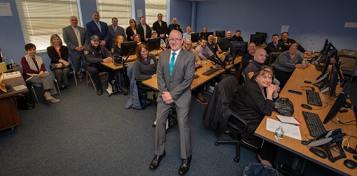 Leadership Coach sits on a desk facing the camera with the park rangers lined up around the room behind him, standing at the walls and working on the computers