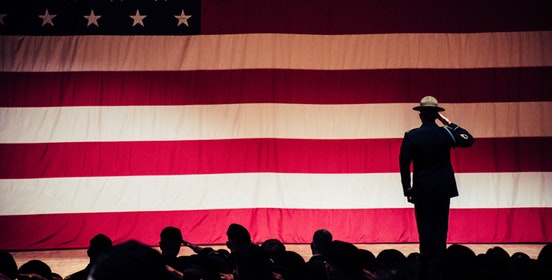 An image of a saluting solider shillouetted against a giant backlit american flag, a shillouetted crowd before him.