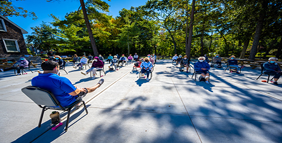 Fitness Instructor, Nick is sitting in a chair facing a group of senior citizens, the group is extending out their leg as part of the workout routine