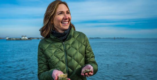 Aileen holds up 2 oysters in her hands while smiling and looking off to camera right, the blue calm waters of the bay rolling behind her.