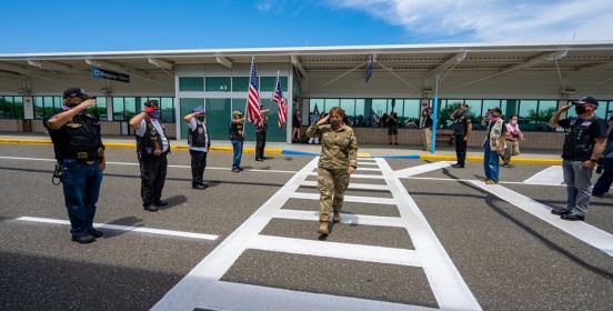 Naval Commander Gardner walks crosswalk out of ISP saluting those who stand with American flag to either side, welcoming her home.