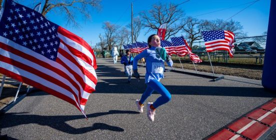 Youth girl in stride crosses finish line as American flags wave on either side of the last stretch.