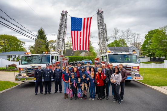 Family and friends of fallen Firefighter Rivelli in group shot under large American flag flown across two FD ladders