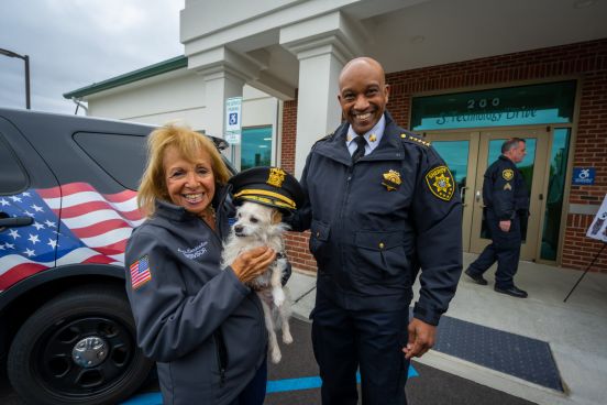 Supervisor Carpenter holds dog beside Sherrif Toulon who holds Sheriff Cap on dog's head.