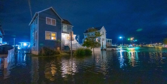 A night image of midnight blue skies and a row of houses illuminated with lights, the flood waters surrounding them up to the doorstep, like a street built on the sea or a community in Venice...dark night time waters reflecting back the sleepy homes' lights in yellow, jade and amethyst ripples.