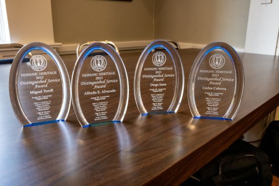 A close-up shot of the 4 glass awards with honoree names etched in on them with lighting shining through over a wood table