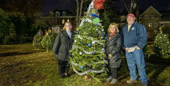Supervisor Carpenter stands with DPW personnel beside a decorated tree with beautifully adorned trees in the distance behind her along the lawn of Brookwood Hall, seen majestic in the background.
