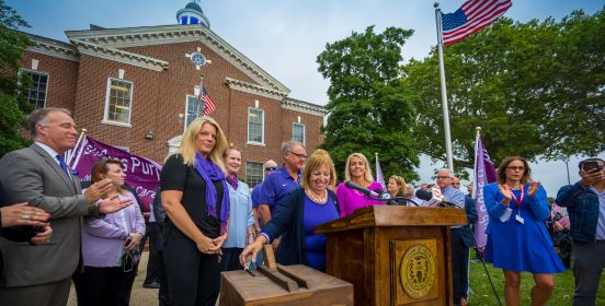 Islip Supervisor, the Islip Town Board, and local officials stand outside the Town Hall building at a podium surroiunded by a crowd as the coupola atop the Town Hall building is illuminated purple
