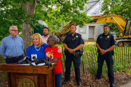 Supervisor at the podium with resident and blighted house prepped for demolition behind her