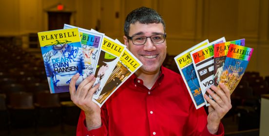  Brian seated in the 401 auditorium holding up an array of playbills, rows of seats in the background behind him.