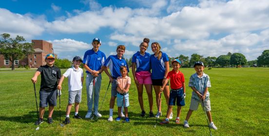 Instructors and kid golfers pose for a photo leaning on clubs over brilliant green fairway and bright blue and cloud speckled skies.
