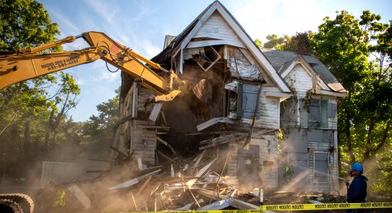 An image capturing the moment the excavator smashes into the zombie house with water from the spray control hoses catching in the morning sun, creating wispy pillars of sunbeams under blue skies.