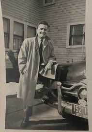 In his prime, Anthony stands with one leg up on vintage car with trench coat collar flipped up