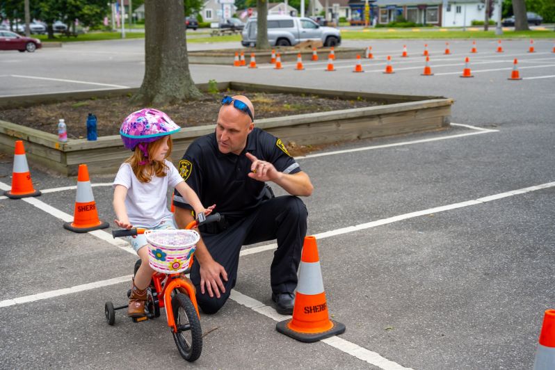 Member of Sheriff's Department gives pointers to youthful girl on tricycle