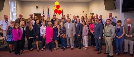 Islip Supervisor Angie Carpenter, members of the Islip Town Board, Food for Hope, and Town of Islip staff pose in the town board room, red and yellow Food for Hope balloons behind them.