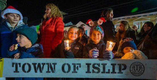Children with smiles and a lit candle look on at the camera above a wooden horse barrier with the words Town of Islip printed on it. 