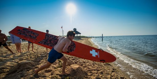 Sun beats down on a lifeguard with red surfboad in his arms as he speeds towards the shoreline, having just begun his trial run.