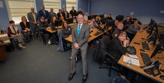 Leadership Coach sits on a desk facing the camera with the park rangers lined up around the room behind him, standing at the walls and working on the computers