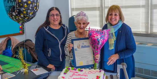  The centenarian stands above her Birthday cake along side Supervisor Carpenter, holding a Town Citation of Recognition and a bouquet of flowers.