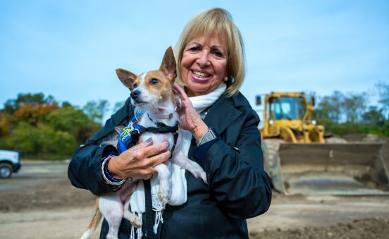 Islip Supervisor Angie Carpenter poses for a photo holding a small pupper, infront of the broken ground where the future shelter will be, a bulldozer in the background.