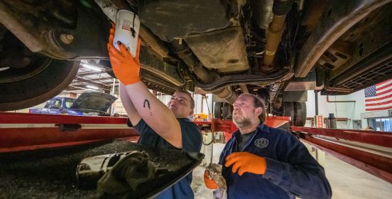 A student holds up a cannister to collect oil as he is taught the process by an experienced technician.