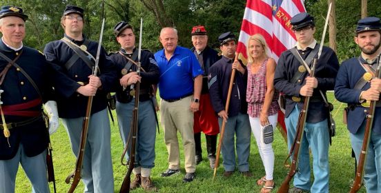 Town Council Members Cochrane and Mullen stand with Town Historian Munkenback and members of the reenactment group on green lawns infront of an american flag.
